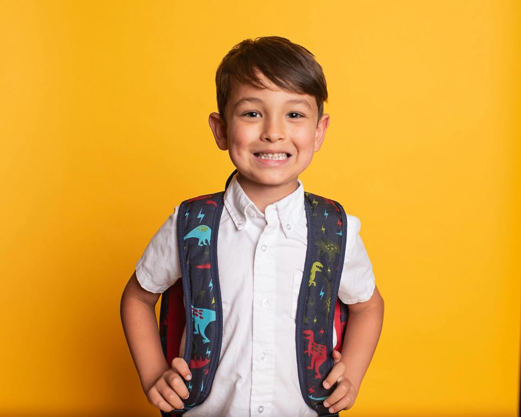 A smiling young child wearing a white shirt and a colorful dinosaur-themed backpack against a bright yellow background.
