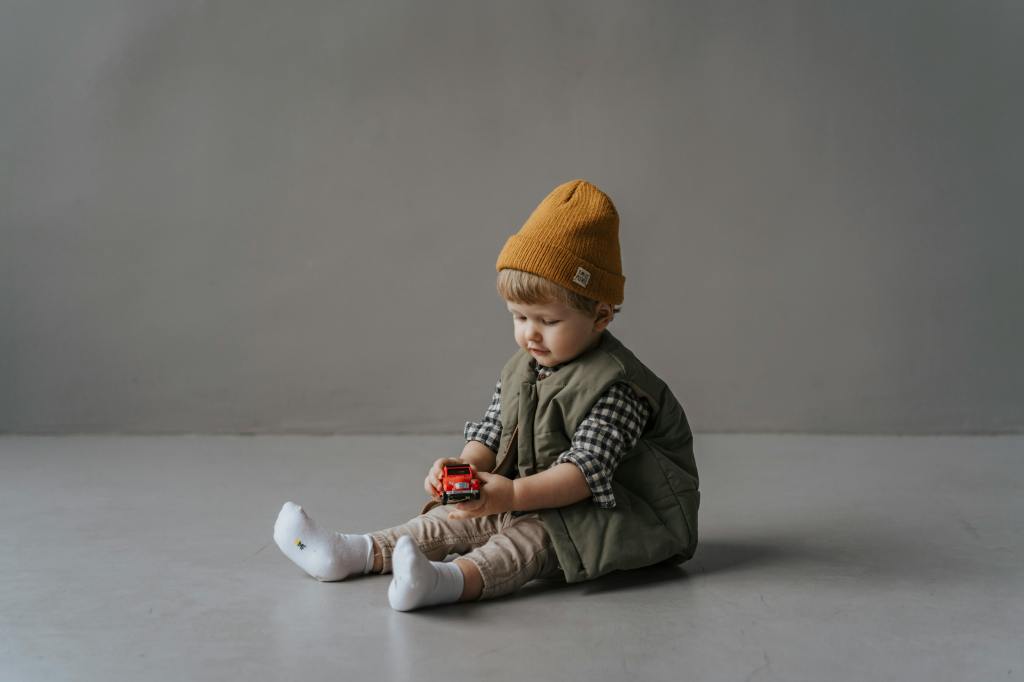 A young child sitting on the floor, playing with a toy car while wearing a mustard-colored beanie, a plaid shirt, and a green vest.