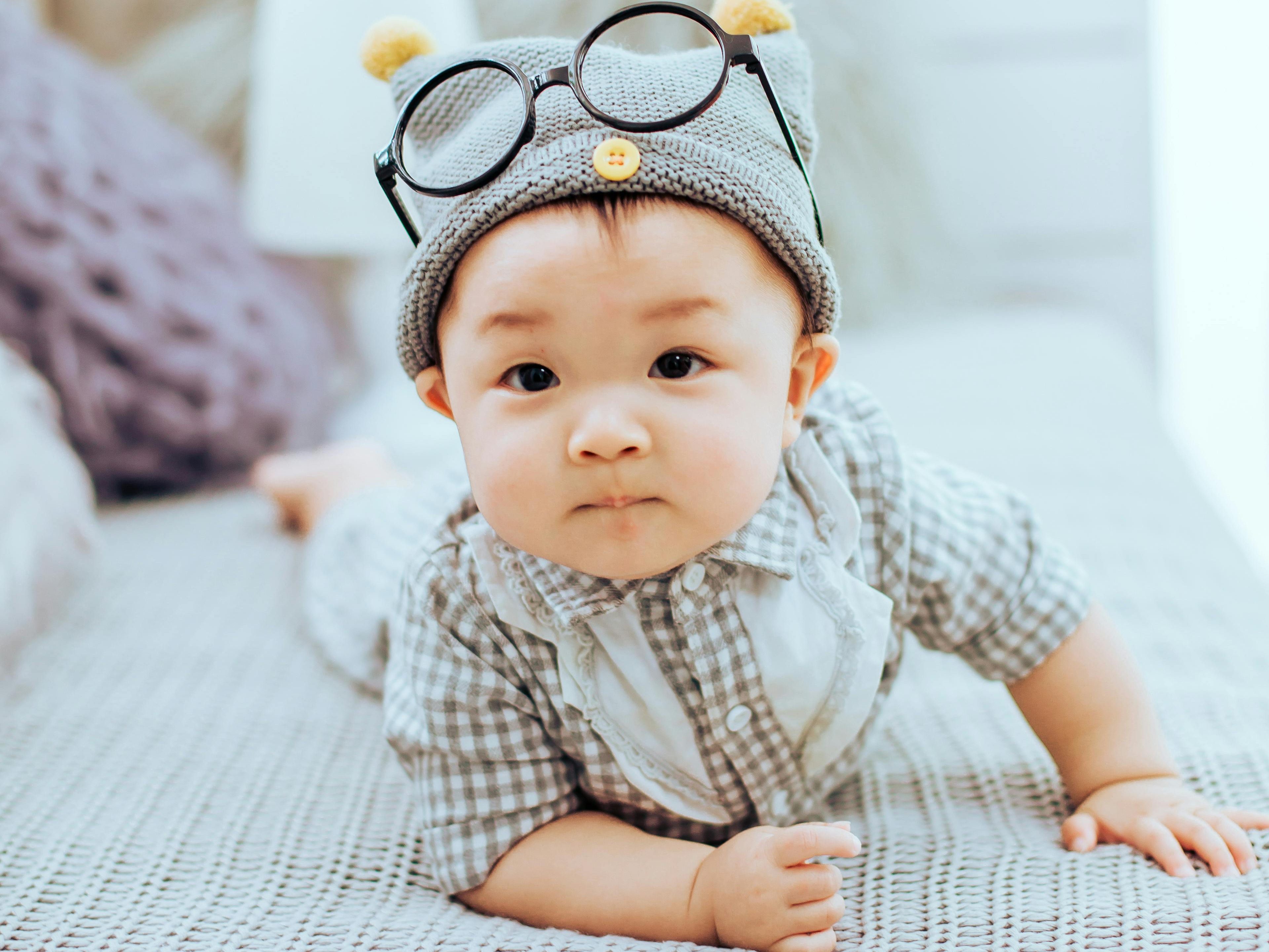 A baby wearing a gray hat with ears and glasses, lying on a bed with a light gray blanket.