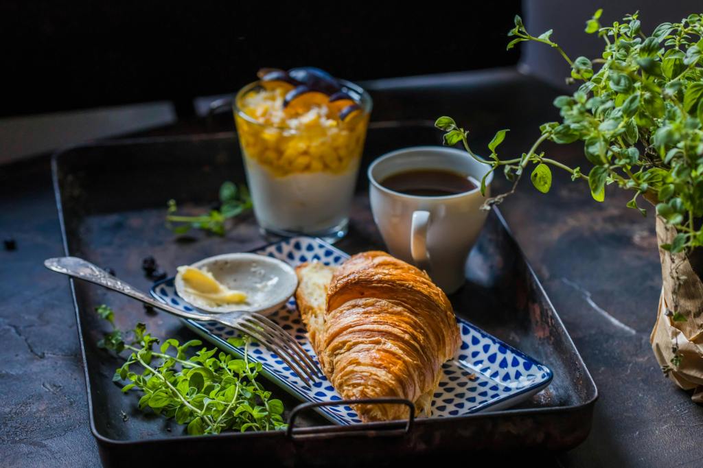 A cozy breakfast tray featuring a croissant with butter, a glass of yogurt with corn, and a cup of coffee, surrounded by fresh herbs.