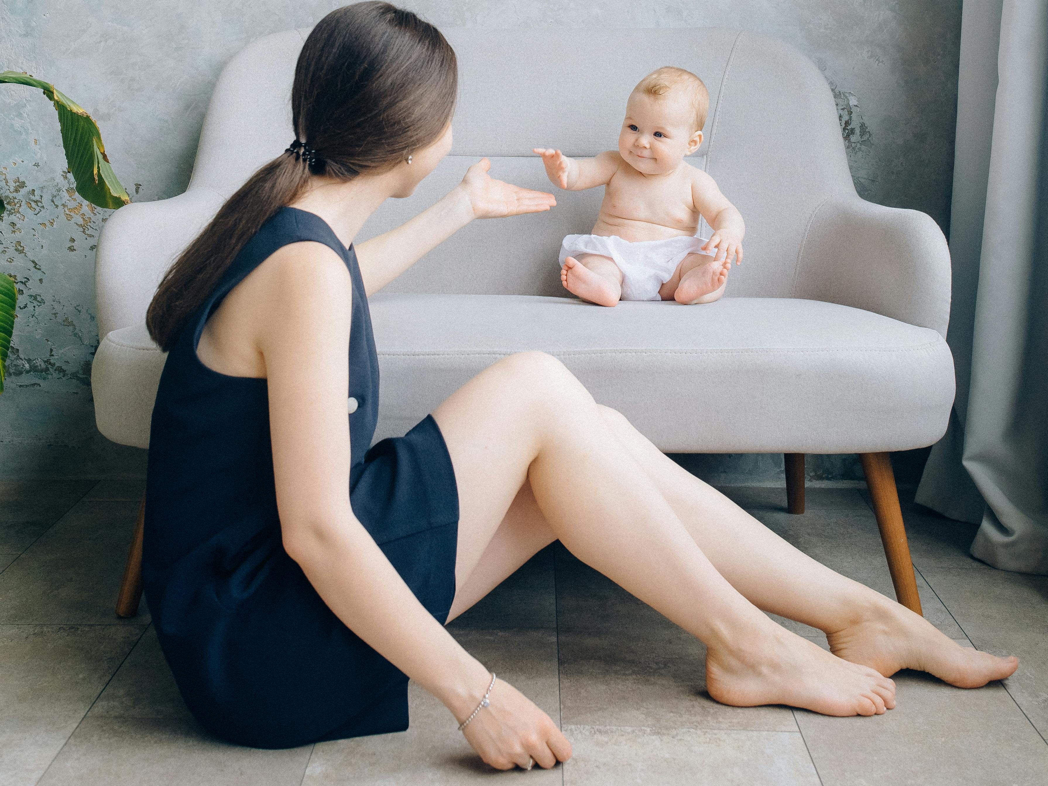 A caregiver interacting with a seated baby, both smiling, in a cozy indoor setting.