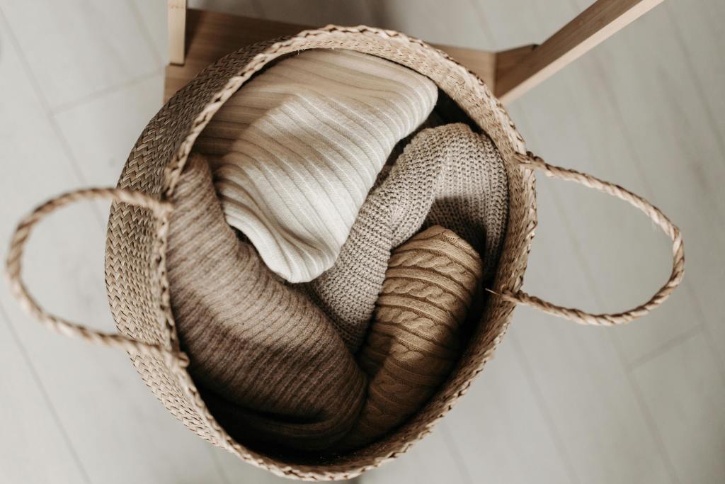 A woven basket filled with neatly rolled soft clothing in neutral tones, including shades of beige, cream, and light brown.