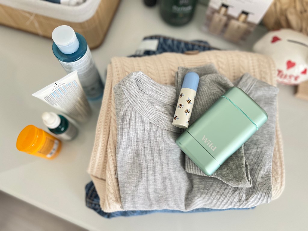 A neatly arranged flat lay of a grey sweater, comfortable pants, and personal care items including a skincare product, a natural deodorant, and hair products. The background features a soft, light-colored surface.
