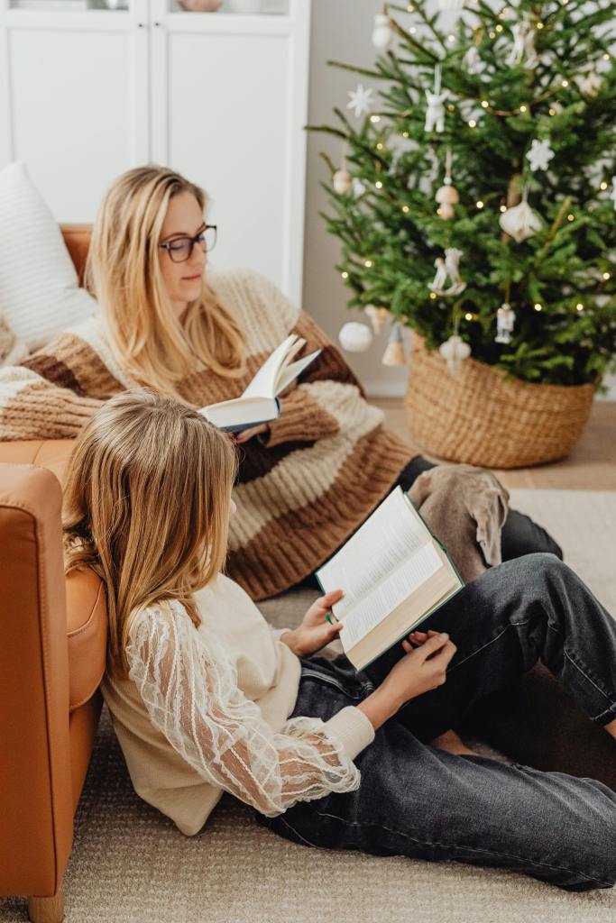 A woman and a child sitting on the floor, both reading books in a cozy living room setting with a Christmas tree in the background.