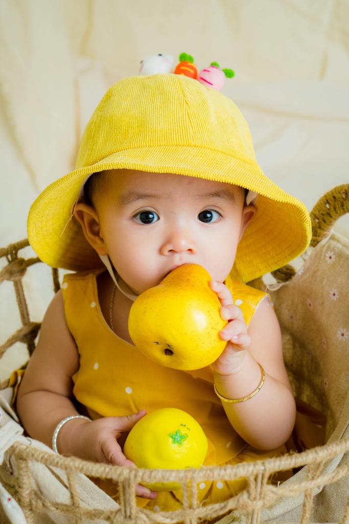 A young child sits in a basket wearing a yellow hat and a yellow dress dotted with white. The child is holding a yellow fruit in one hand and a lemon in the other, looking curiously at the camera.
