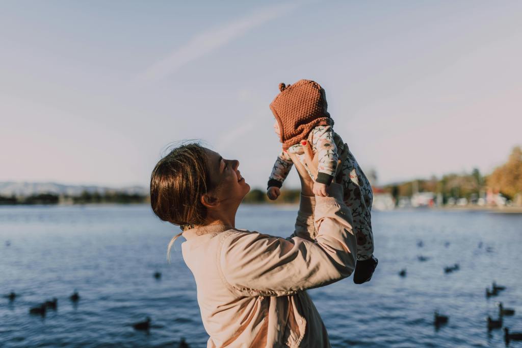 A parent joyfully holds a baby up in the air by a lake, surrounded by water and ducks, under a clear sky.