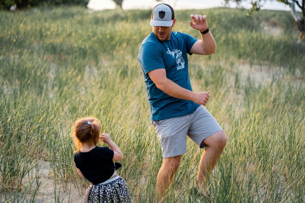 A father and young daughter play together in a grassy field, with the father dancing or making playful movements while the daughter watches, smiling and engaged.
