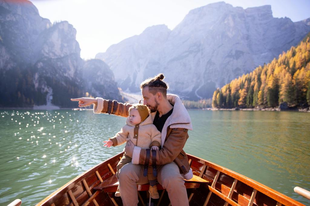 A father sitting in a boat by a lake, holding a baby and pointing towards the mountains in the distance.