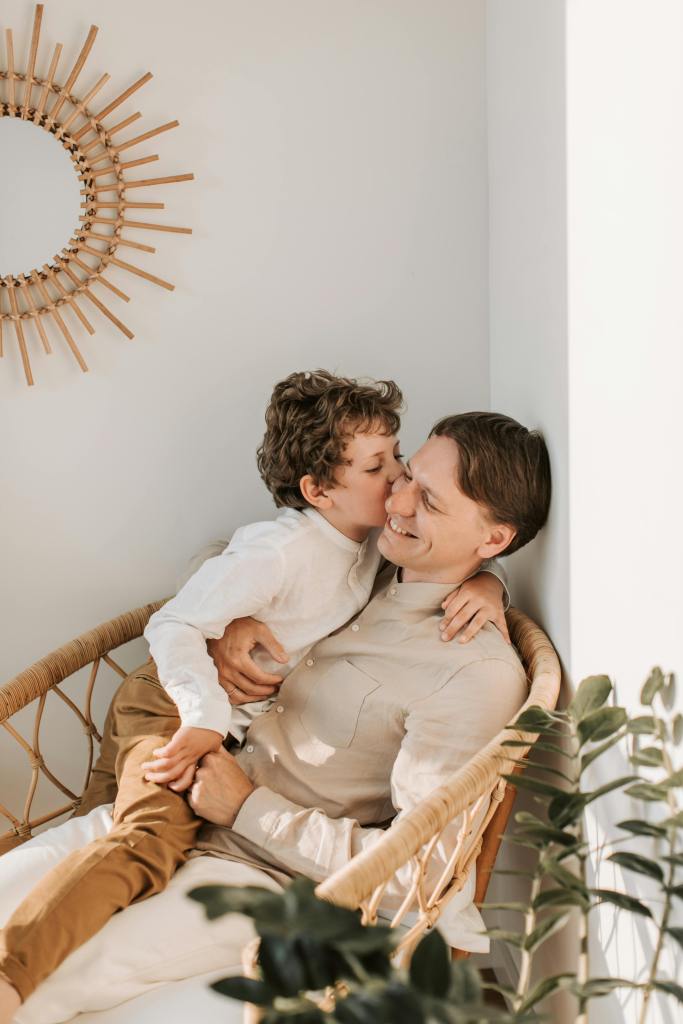 A father and child sharing a joyful moment, with the child giving the father a kiss while sitting together in a cozy chair surrounded by plants and a decorative mirror.