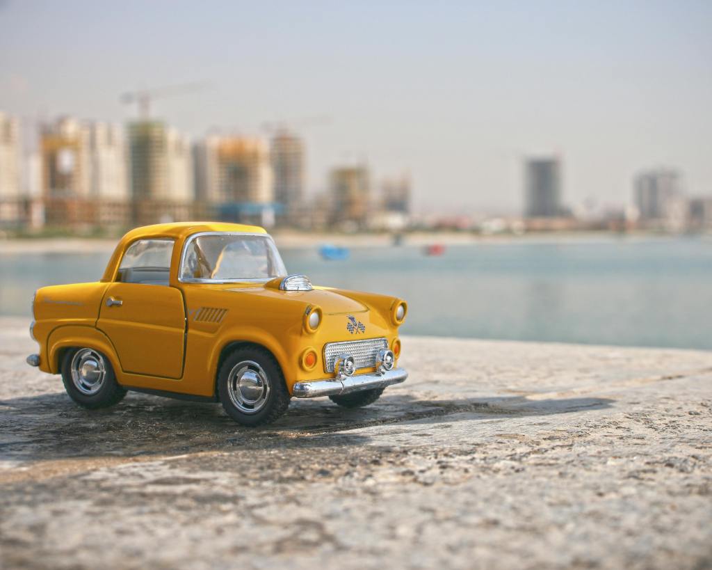 A yellow toy car is parked on a stone surface with a blurred city skyline and water in the background.
