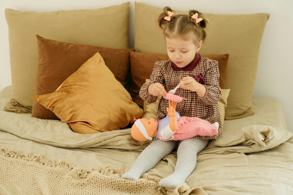 A young girl with pigtails is sitting on a bed, dressed in a checkered dress, playing with a doll. She is holding a spoon and appears to be feeding the doll while surrounded by soft cushions.