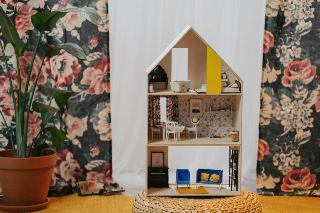 A modern dollhouse with detailed interiors, set against a patterned background of flowers and foliage.