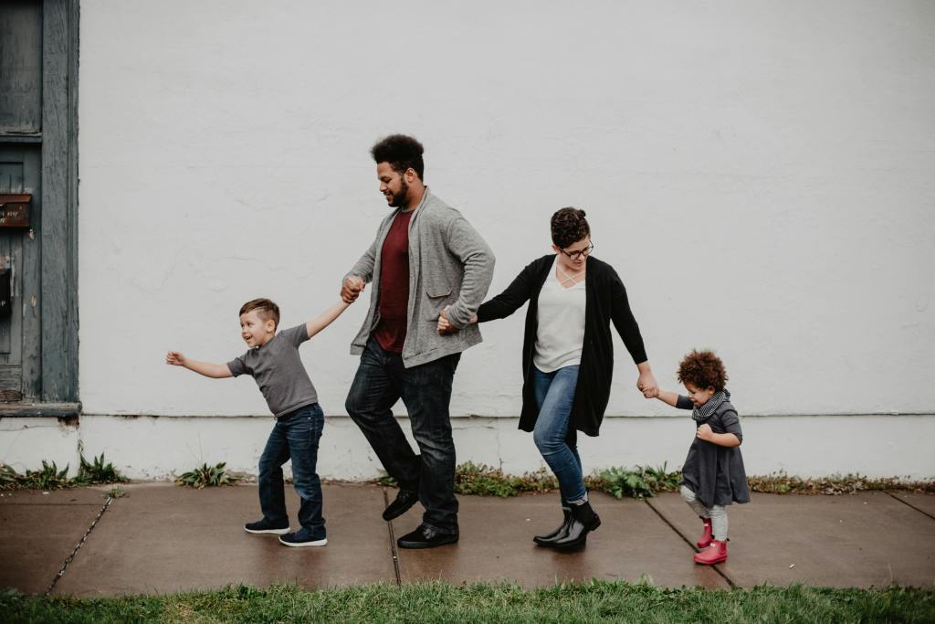 A family of four, including two adults and two children, walking hand in hand along a sidewalk in front of a white wall, with greenery in the foreground.