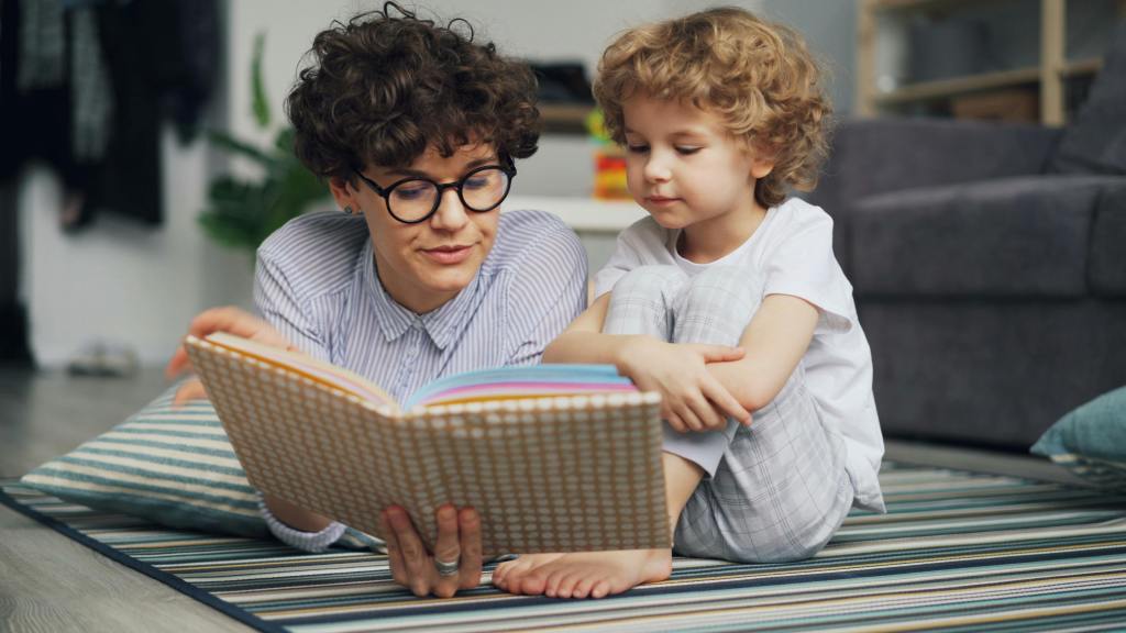A parent and a child lying on a rug, reading a book together in a cozy, modern living space.
