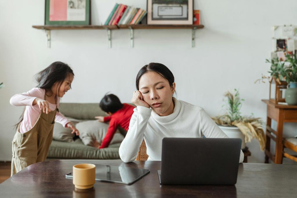 A stressed parent working on a laptop at home while two children play in the background, one of them playfully interacting with the parent.