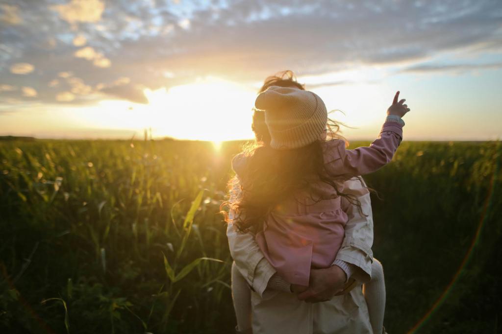 A parent holding a child on their shoulder, both gazing towards the sunset in a field, capturing a moment of connection and tranquility.